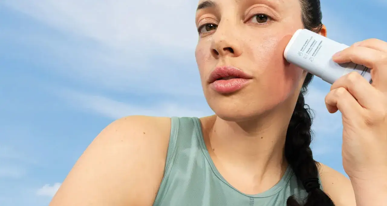 Une femme applique un &eacute;cran solaire physique sur sa joue, sous un ciel bleu.