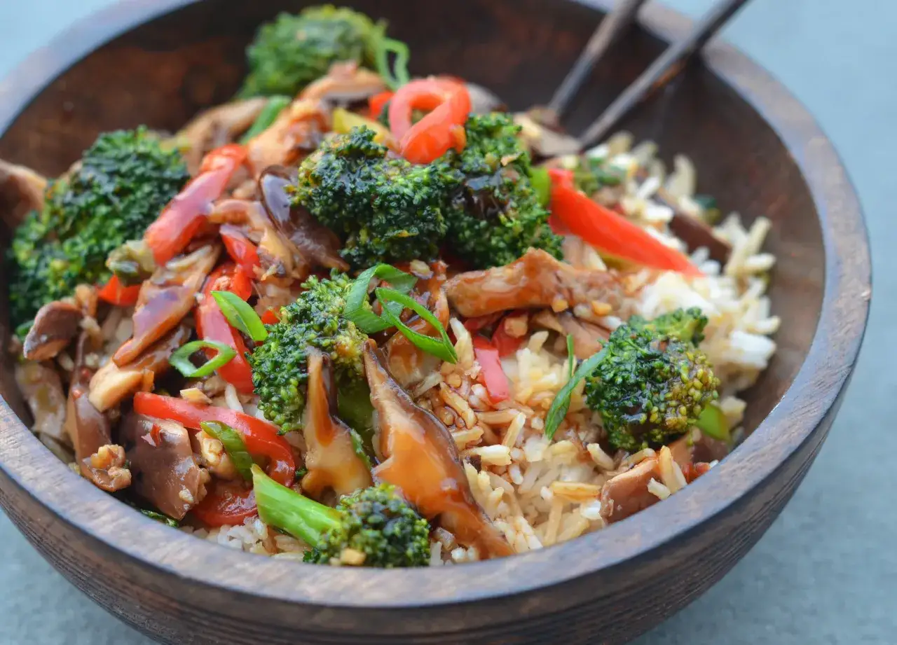 Un bol en bois rempli d'un savoureux poêlée de légumes avec du riz. Brocolis, poivrons rouges et champignons se mélangent dans une sauce appétissante.