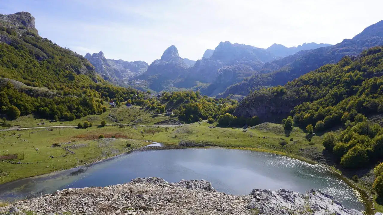 A serene lake reflects the majestic mountains and lush green slopes dotted with houses, perfect for a Montenegro itinerary.