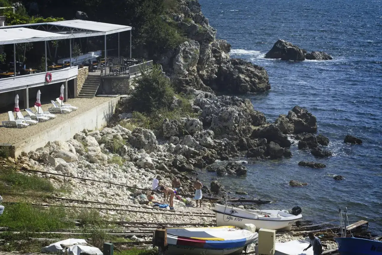 Słoneczny dzień na plaży w Bułgarii. Ludzie odpoczywają przy morzu, obok łodzie i restauracja na klifie. Idealne miejsce na wakacje z elementami **camping bułgaria**.