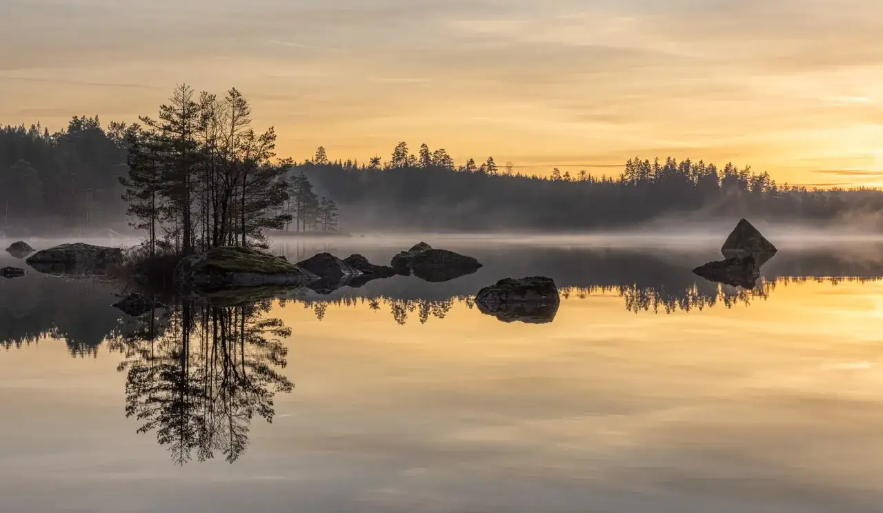 Tidig morgon vid en sjö i Kilsbergen. Träd och stenar speglar sig i det stilla vattnet under en gyllene soluppgång. En perfekt plats för kilsbergen vandring.