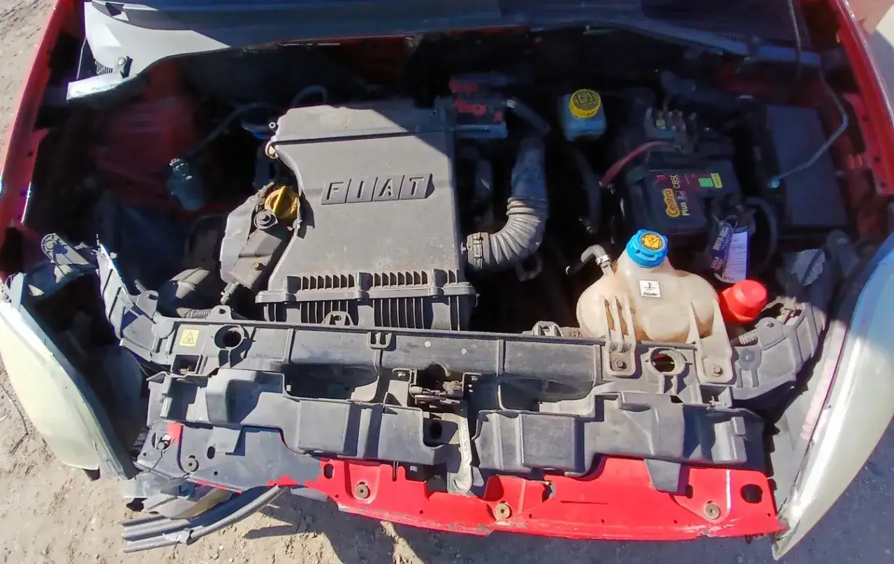 Silnik fiat grande punto 1.4. Widoczny jest plastikowy element z napisem FIAT, akumulator, zbiorniczek płynu chłodniczego i inne elementy komory silnika.