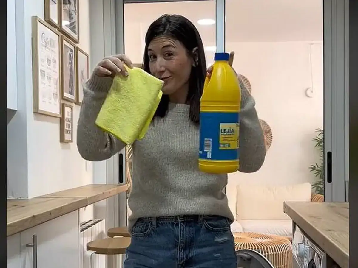 Mujer sonriente con un pa&ntilde;o amarillo y una botella de lej&iacute;a, lista para limpiar la lavadora.