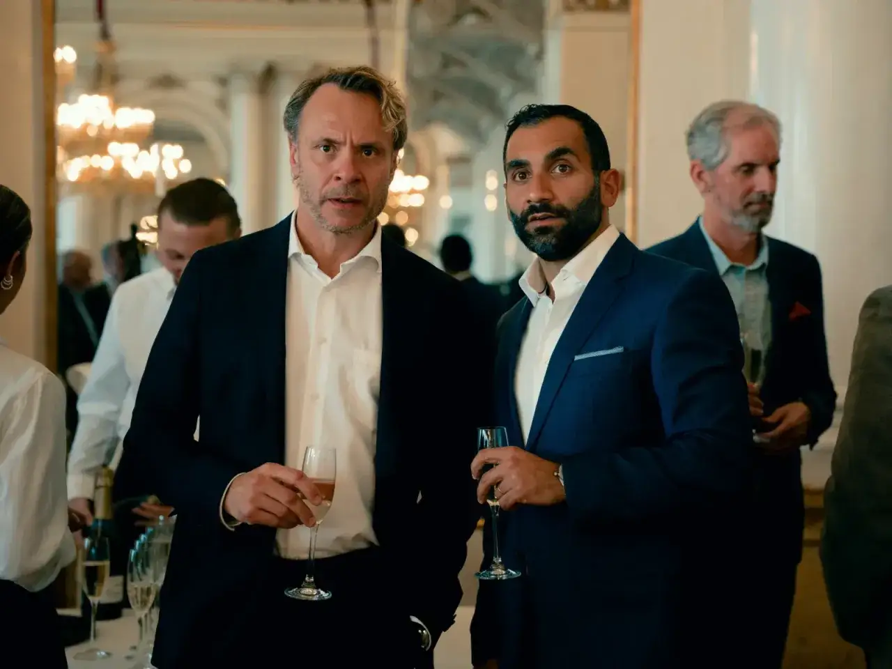 Två män i kostym med champagneglas i handen, ser ut som en del av rollistan i hundarna.