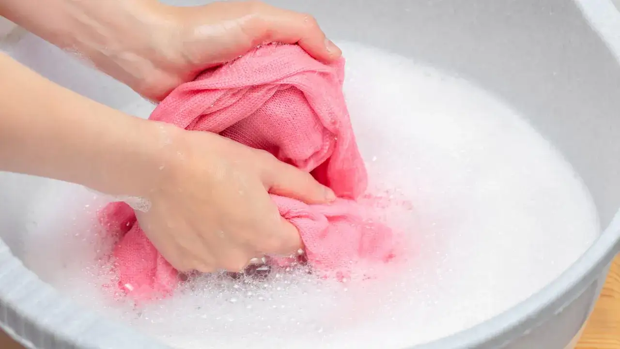 Hände waschen einen rosafarbenen Pullover in schaumigem Wasser. Ein sanfter Wollprogramm-Waschgang ist ideal für solche empfindlichen Textilien.