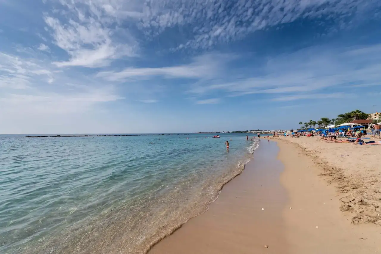 Słoneczna plaża z turkusową wodą i ludźmi relaksującymi się na piasku. Idealne miejsce na wakacje w hotelu Leptos Paphos Gardens Holiday Resort.