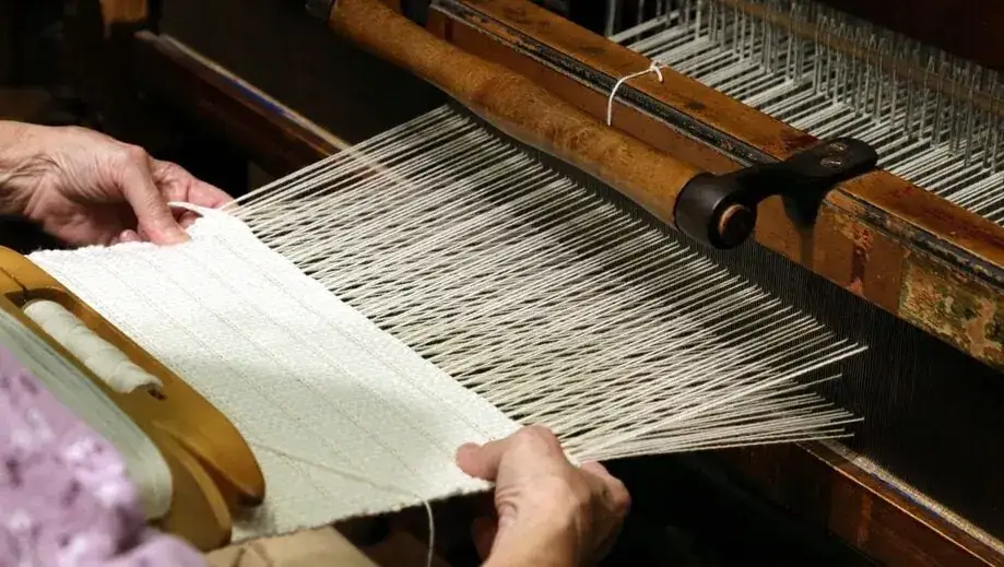 Mains habiles travaillant sur un m&eacute;tier &agrave; tisser ancien, cr&eacute;ant un tissu blanc. La fabrication textile, un art ancestral.