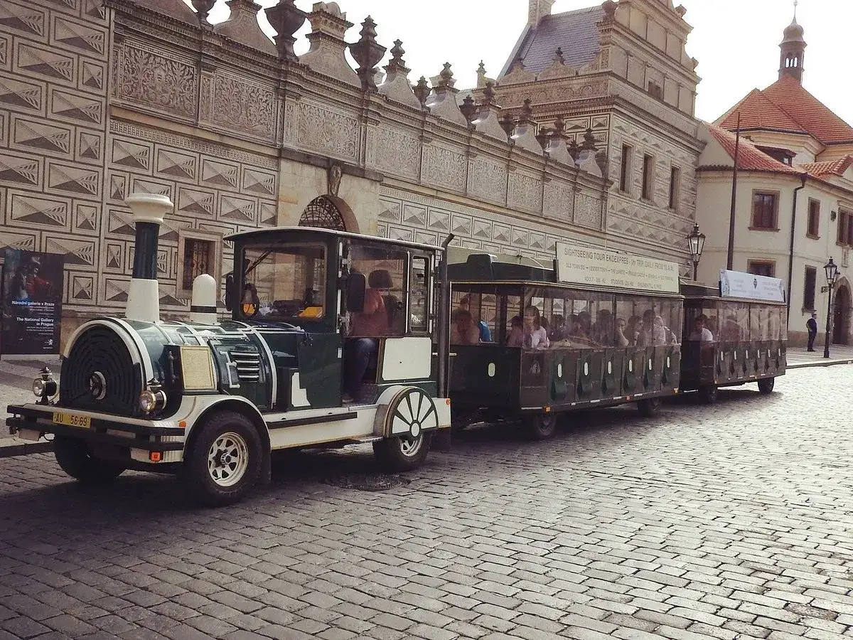 A tourist train, perfect for day trips from Prague by train, waits on a cobblestone street in front of a historic building.
