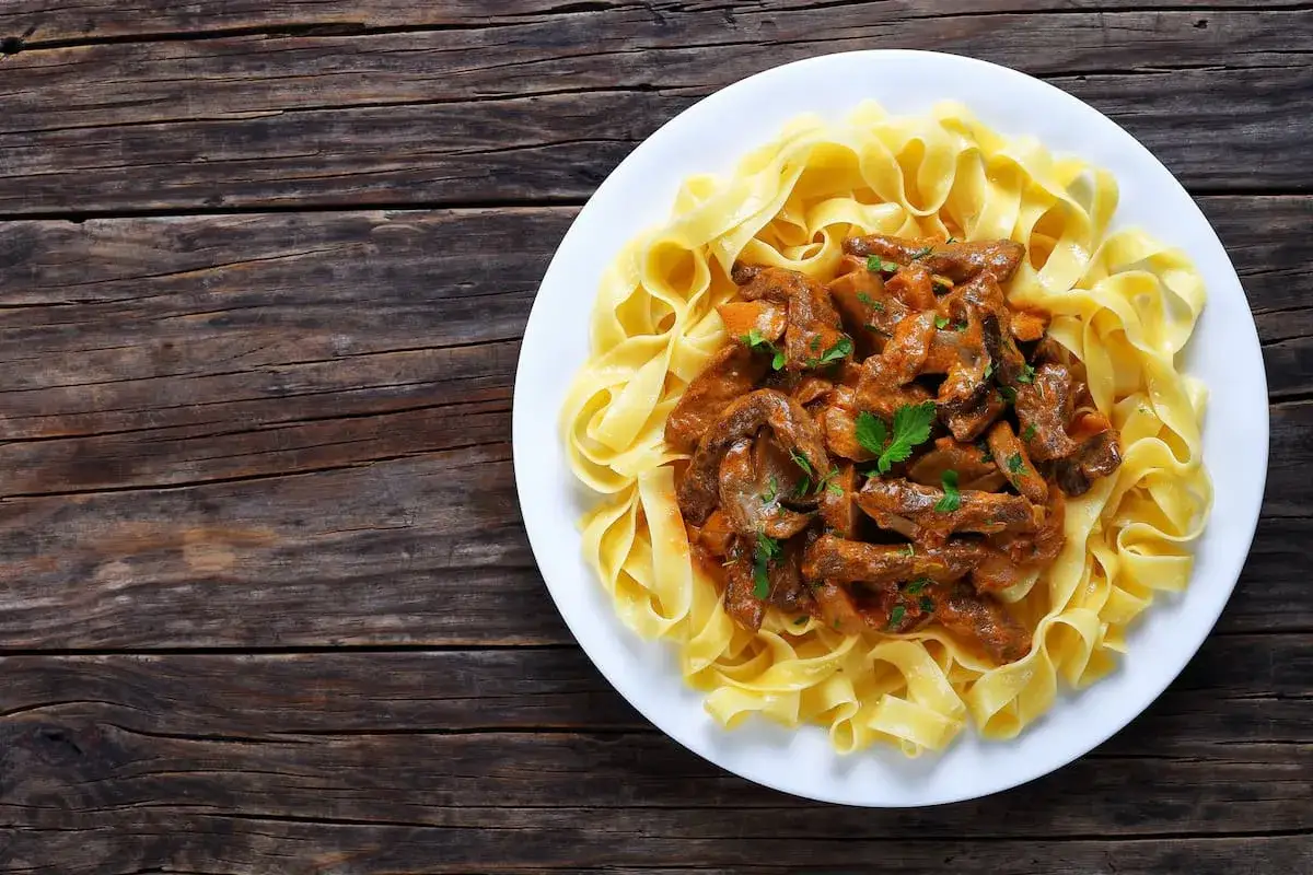 Un plato de fideos con carne, cubierto con una rica salsa cremosa y perejil fresco, servido sobre una mesa de madera r&uacute;stica.