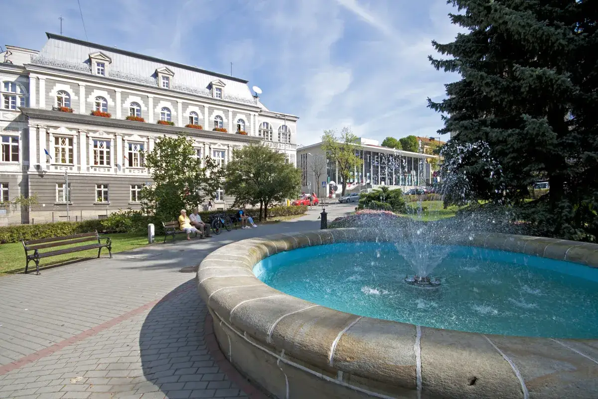 Słoneczny dzień na rynku w Tarnowie. Fontanna w centrum, obok ławki z odpoczywającymi ludźmi i zabytkowy budynek.