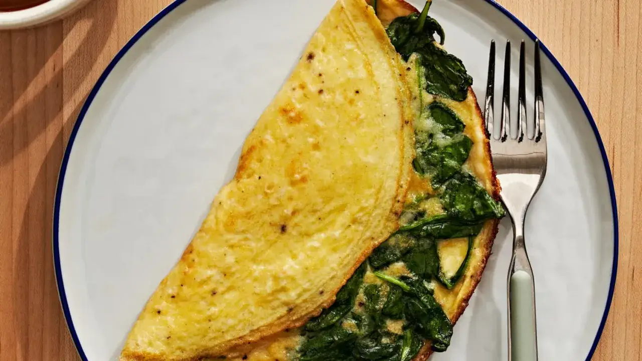 Złożony omlet bez ubijania z zielonym szpinakiem i serem, podany na białym talerzu z niebieską obwódką. Obok leży widelec.
