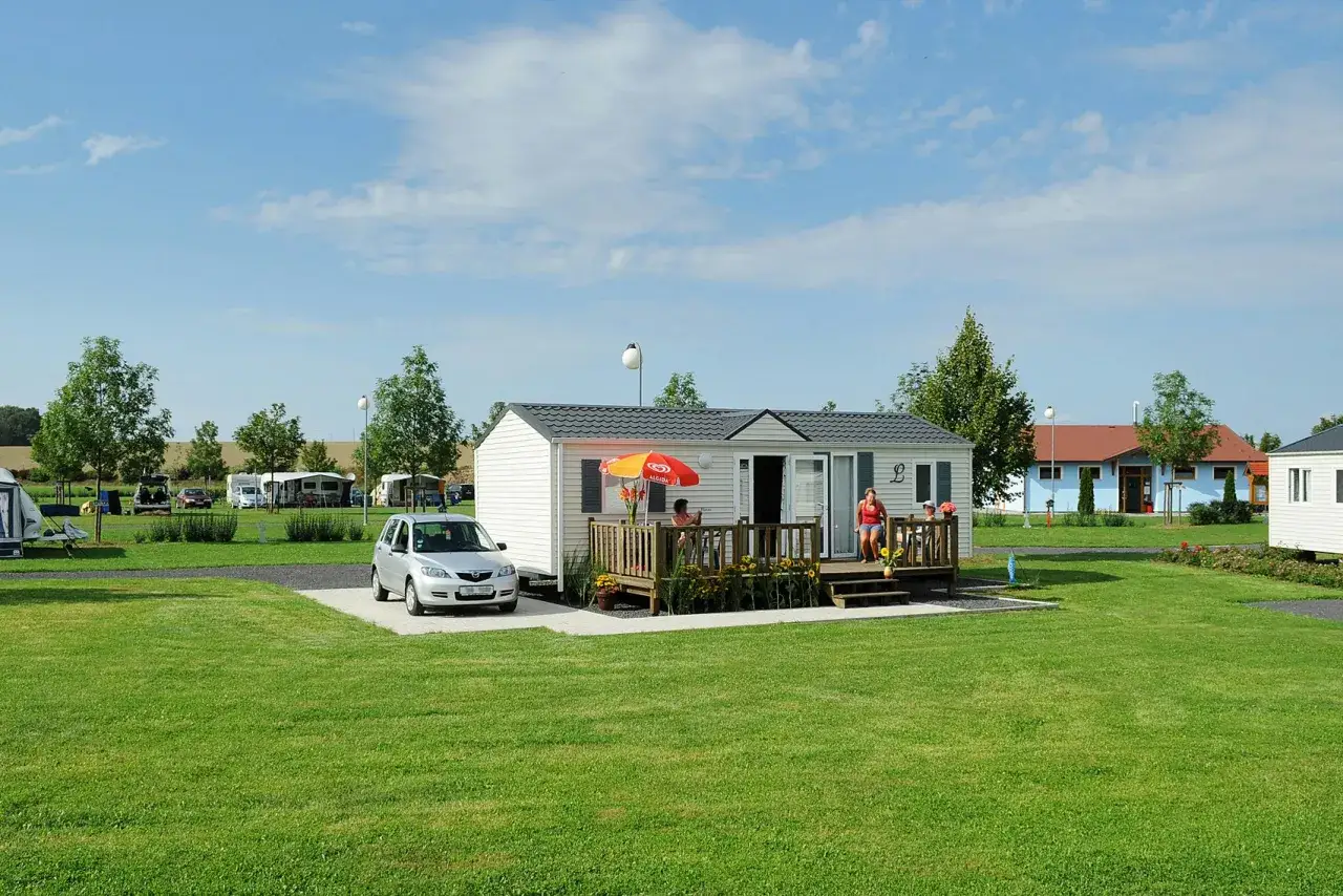 Ein sonniger Tag auf dem Campingplatz Vaclav. Ein Wohnwagen steht neben einem Mobilheim mit Terrasse, auf der zwei Frauen sitzen. Ein Auto parkt davor.