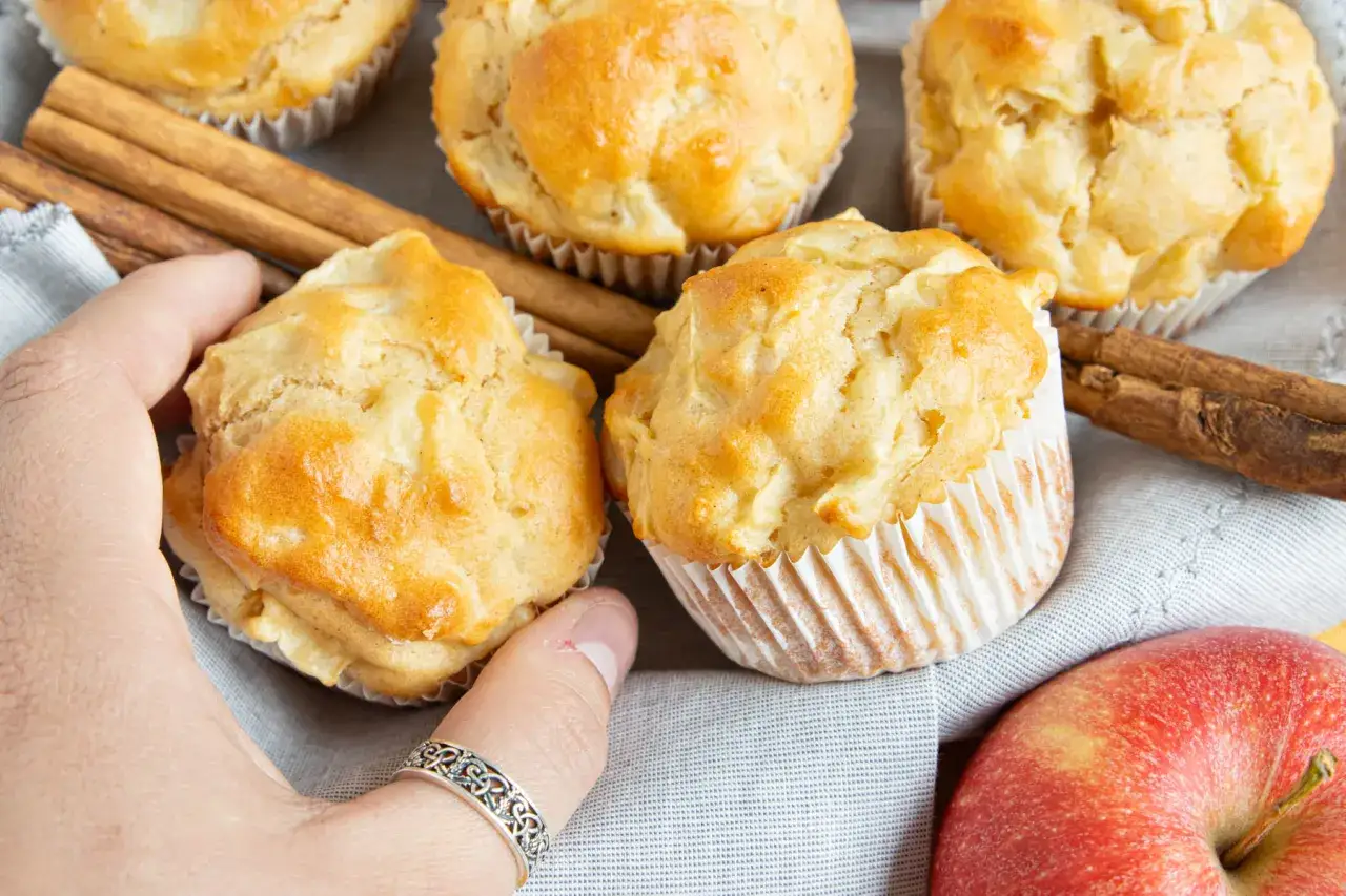 Hand greift nach saftigen apfelmuffins ohne zucker, daneben Zimtstangen und ein roter Apfel.