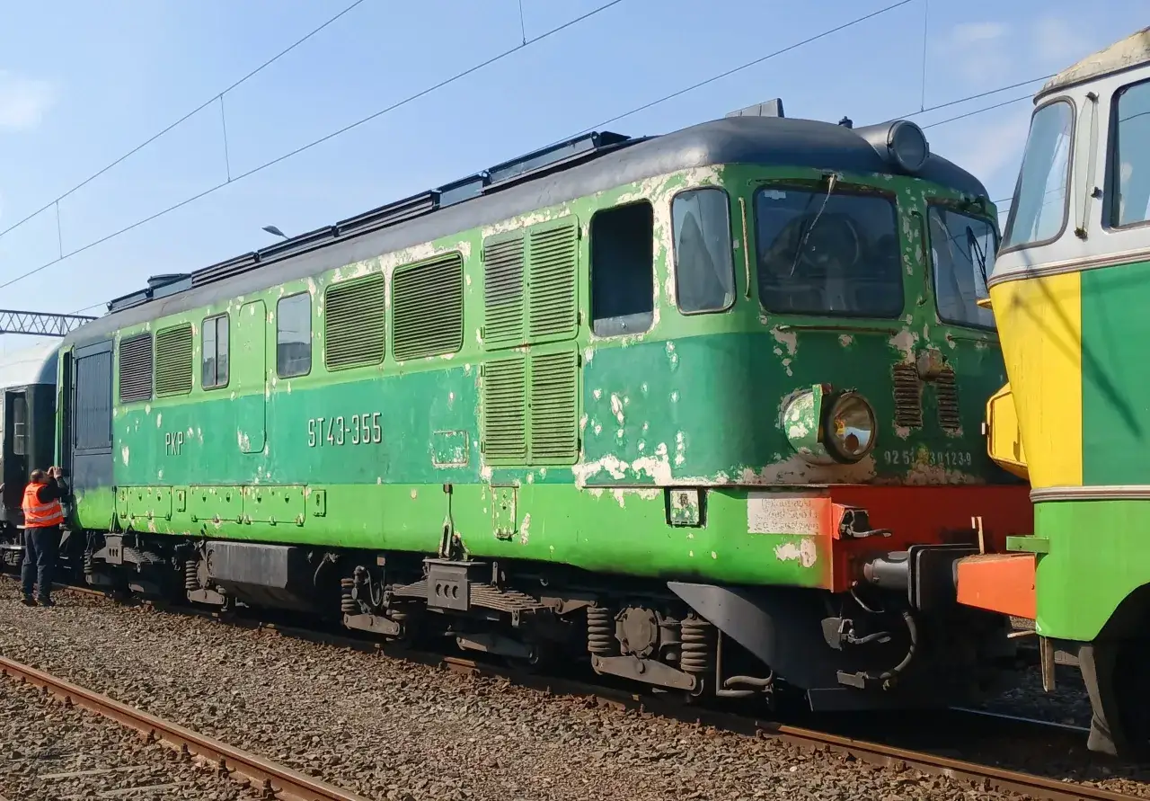 Zielona lokomotywa ST43-355 PKP z odrapaną farbą stoi na torach obok innej maszyny.