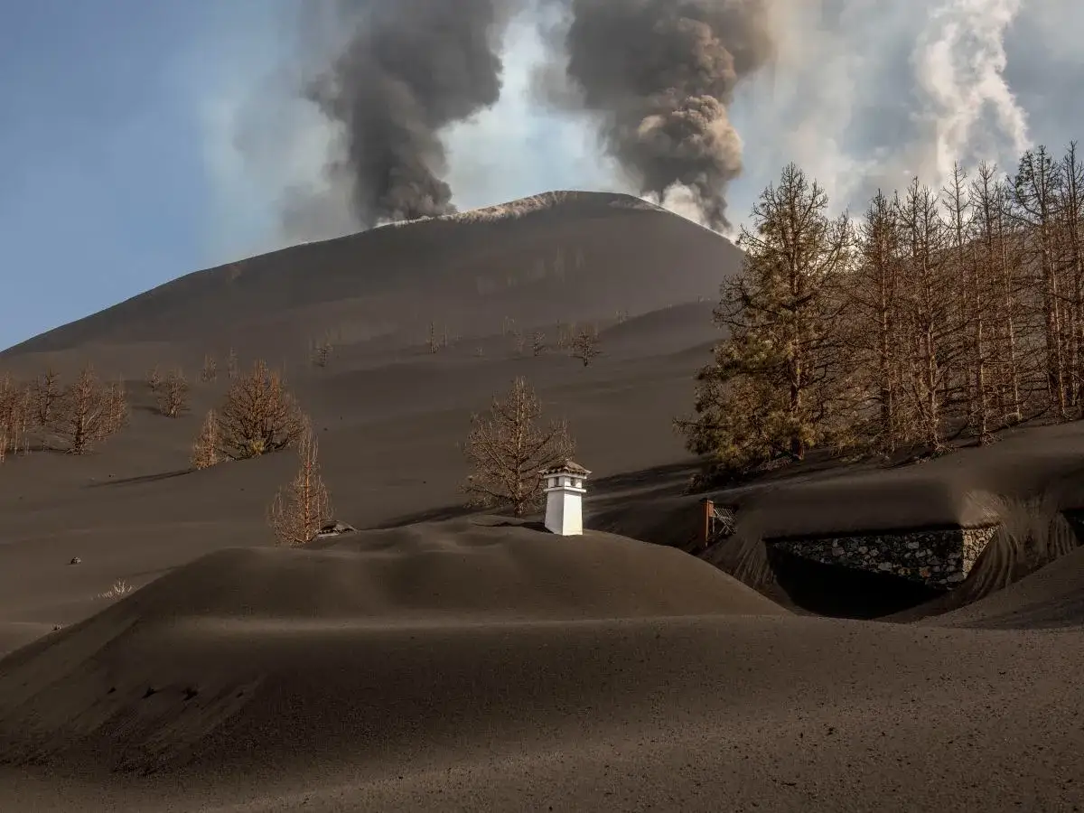 Popiół wulkaniczny pokrywa krajobraz La Palma. W oddali widać dymiący wulkan, a na pierwszym planie mały biały komin. Obraz przypomina o zagrożeniu tsunami La Palma.