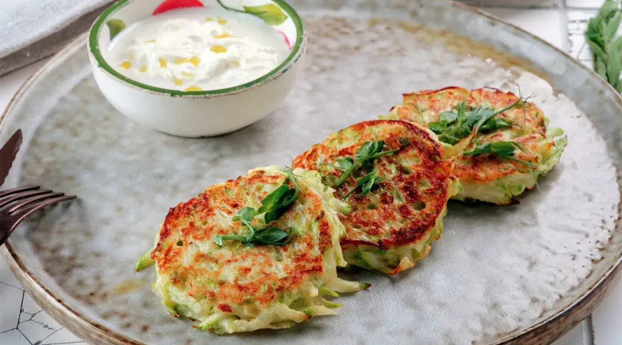 Drei goldbraune Kartoffel-Zucchini-Puffer mit frischen Sprossen garniert, daneben eine Sch&uuml;ssel cremiger Dip.