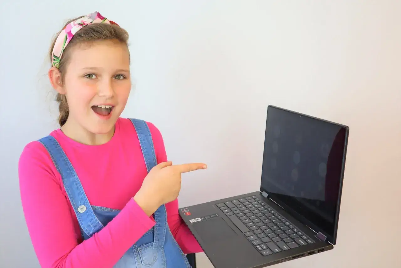Uśmiechnięta dziewczynka w różowej bluzce i ogrodniczkach wskazuje palcem na ekran laptopa, na którym widać ikony rozmów. To jej przygoda z Helen Doron stream.