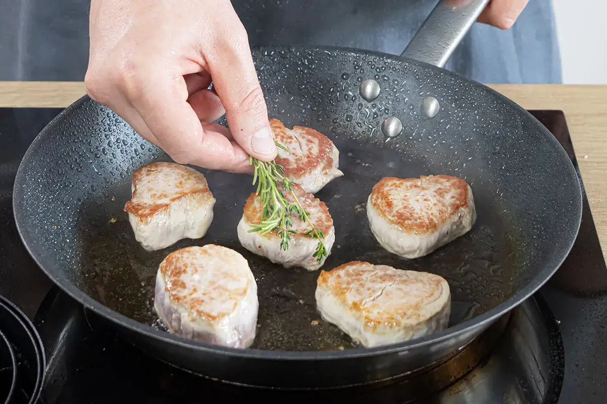 Zarte schweine medaillons braten in der Pfanne, eine Hand f&uuml;gt frischen Rosmarin hinzu.