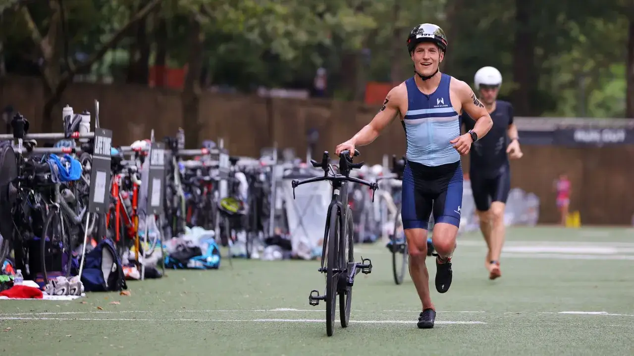 Läufer mit Rennrad beim olympischen Triathlon. Im Hintergrund weitere Athleten und Fahrräder.