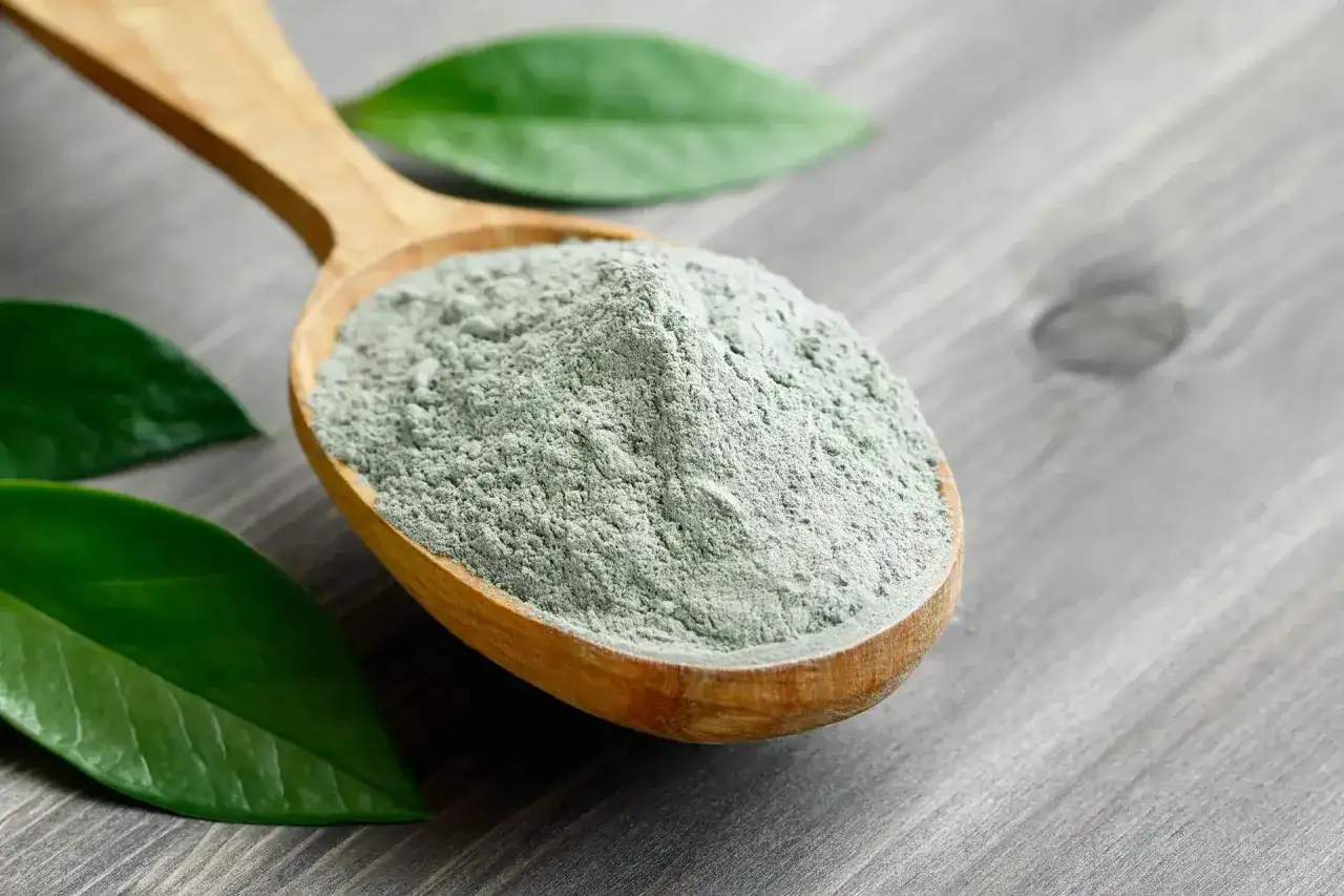 Argilla verde a cosa serve? Questa polvere finissima su un cucchiaio di legno, circondata da foglie verdi, è un tesoro per la pelle.