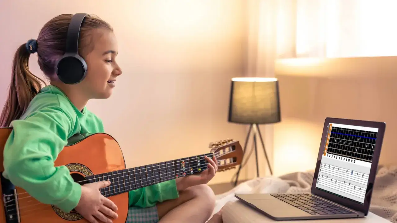 Dziewczynka w słuchawkach uczy się grać na gitarze, korzystając z lekcji online. Na ekranie laptopa widać tabulaturę, idealną do nauki piosenek na gitarę dla początkujących dzieci.