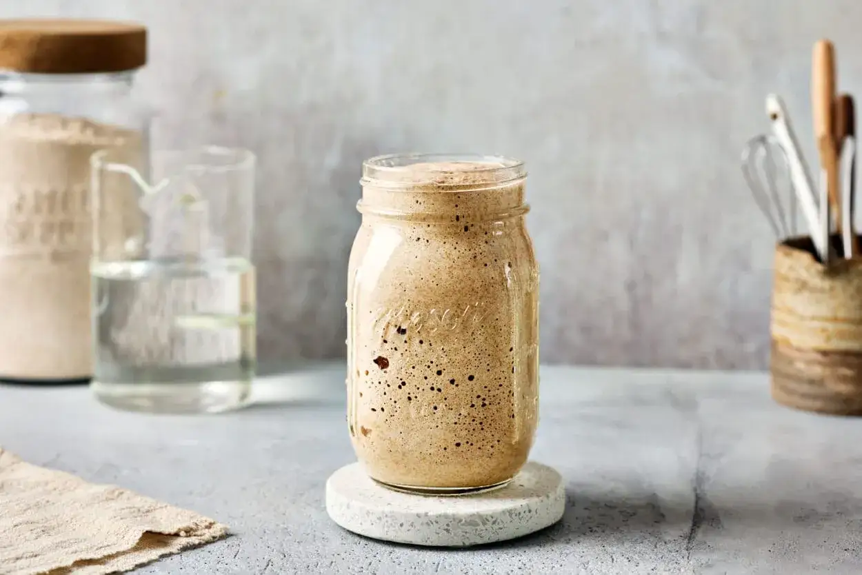 Ein Glas mit sprudelndem Sauerteig-Starter, angesetzt mit Dinkelmehl. Daneben ein Glas Wasser und ein Behälter mit Küchenutensilien.