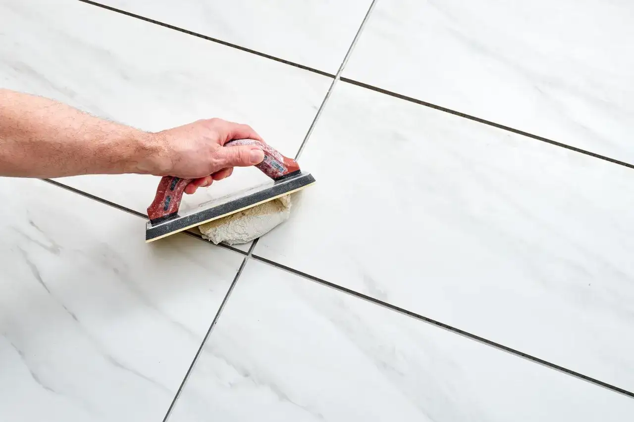 A hand uses a grout float to apply grout to the seams between white marble-look porcelain tiles, showcasing the best grout for porcelain tile.