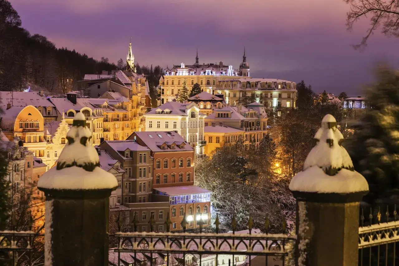 Winterliche Pracht in Karlsbad: verschneite Geb&auml;ude unter einem lila Himmel, beleuchtete Fenster und B&auml;ume.