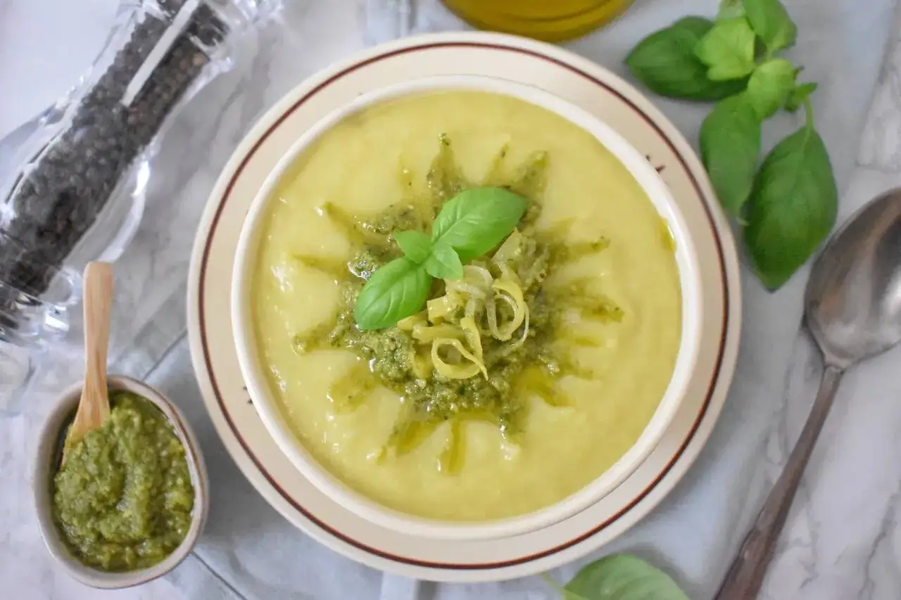 Kremowa zupa porowa z serkiem topionym, udekorowana pesto i listkiem bazylii. Obok miseczka z pesto i młynek do pieprzu.