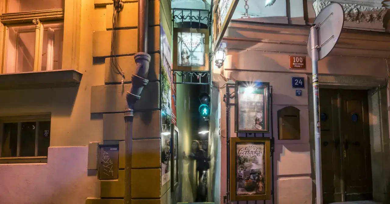 Wąska uliczka w Pradze, gdzie światło sygnalizacji świetlnej oświetla przejście. Widać szyld restauracji 