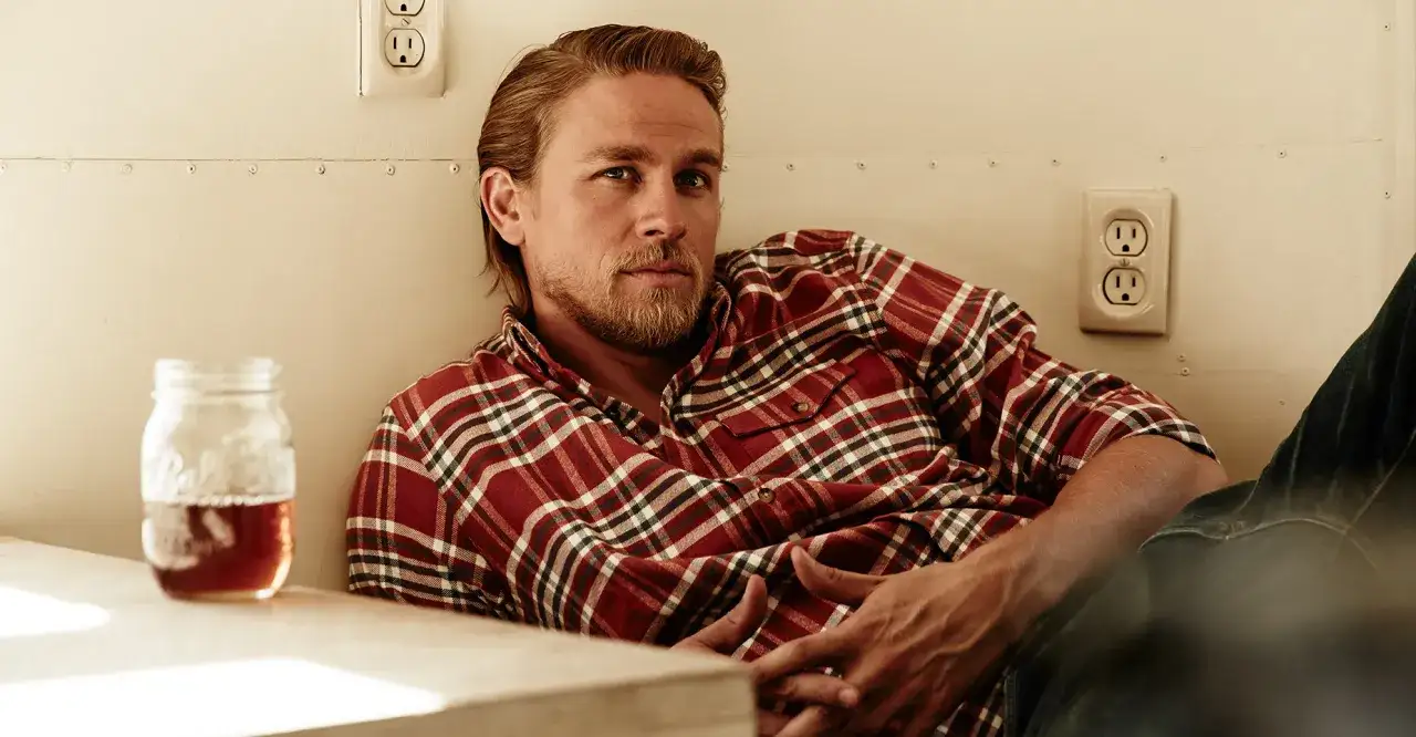 Charlie Hunnam i rödrutig skjorta lutar sig mot en vägg med en burk med dryck bredvid sig.