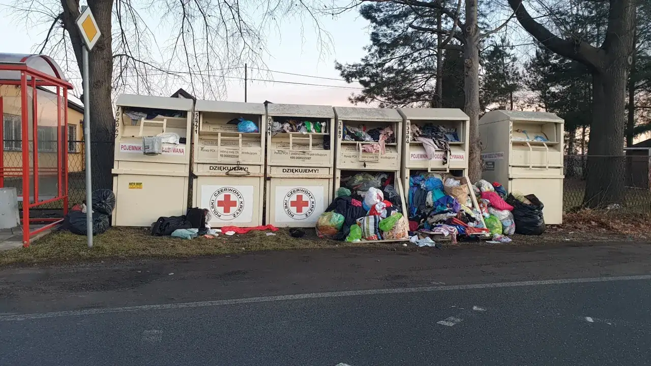 Pojemniki na odzież PCK, gdzie oddać dobre ubrania. Obok kontenerów leżą worki i ubrania.