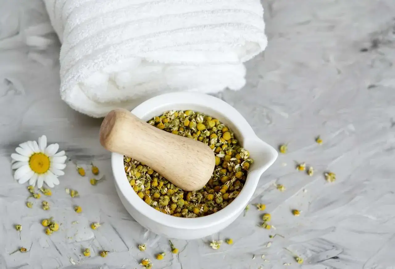 Mortaietto con camomilla secca, ideale per trattamenti naturali per capelli. Un fiore di camomilla e un asciugamano bianco completano la scena.