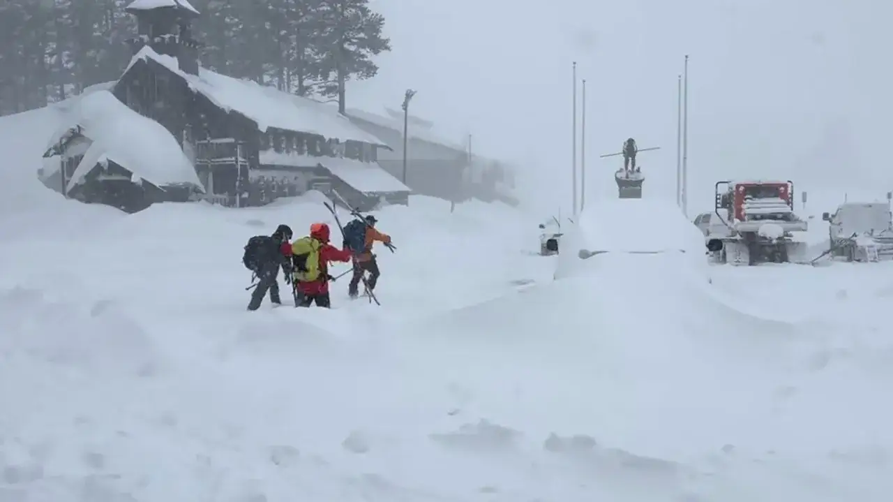 Tres esquiadores avanzan entre la nieve, cerca de un edificio cubierto y vehículos sepultados. Las **dos avalanchas** han transformado el paisaje en un desafío blanco.
