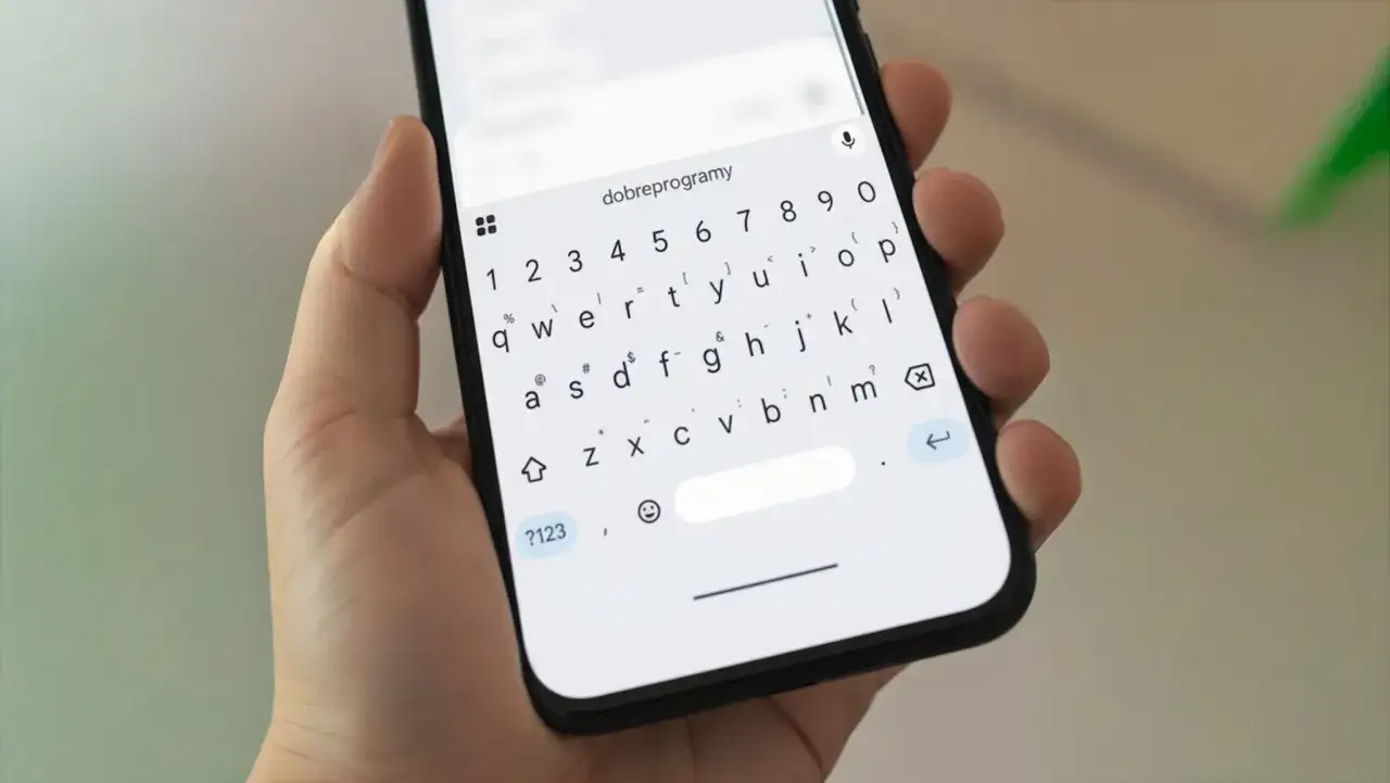 Dłoń trzyma smartfon z klawiaturą ekranową. Dowiedz się, jak napisać pierwiastek na telefonie, korzystając z symboli specjalnych.