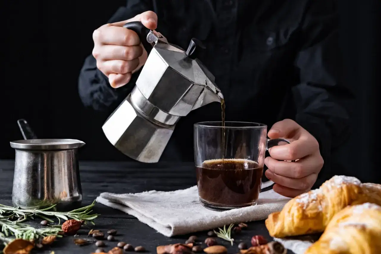 Cafetière italienne inox noircie versant un café chaud dans une tasse, avec des croissants et des épices.