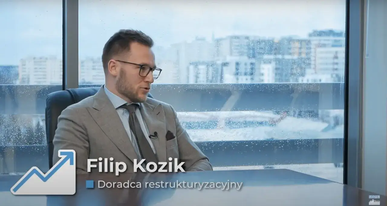 Mężczyzna w garniturze siedzi przy biurku, za nim okno z widokiem na miasto.