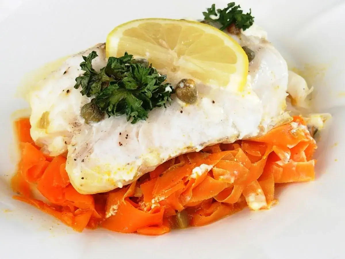 Délicieux cabillaud en papillote, servi sur des carottes râpées, garni de persil, câpres et d'une tranche de citron.