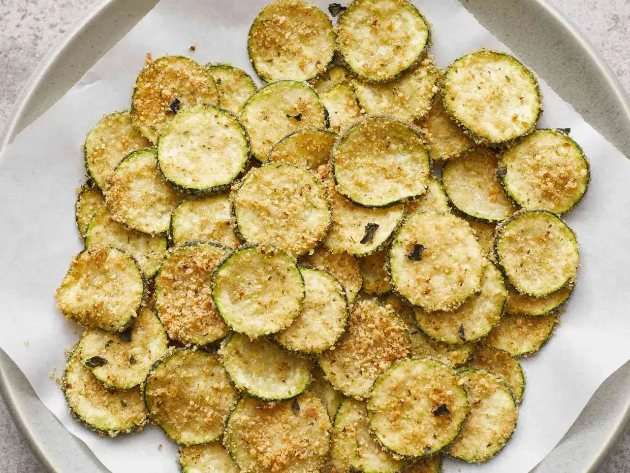 Knusprige Zucchini-Chips, goldbraun gebacken und mit Kr&auml;utern bestreut, liegen auf Backpapier.