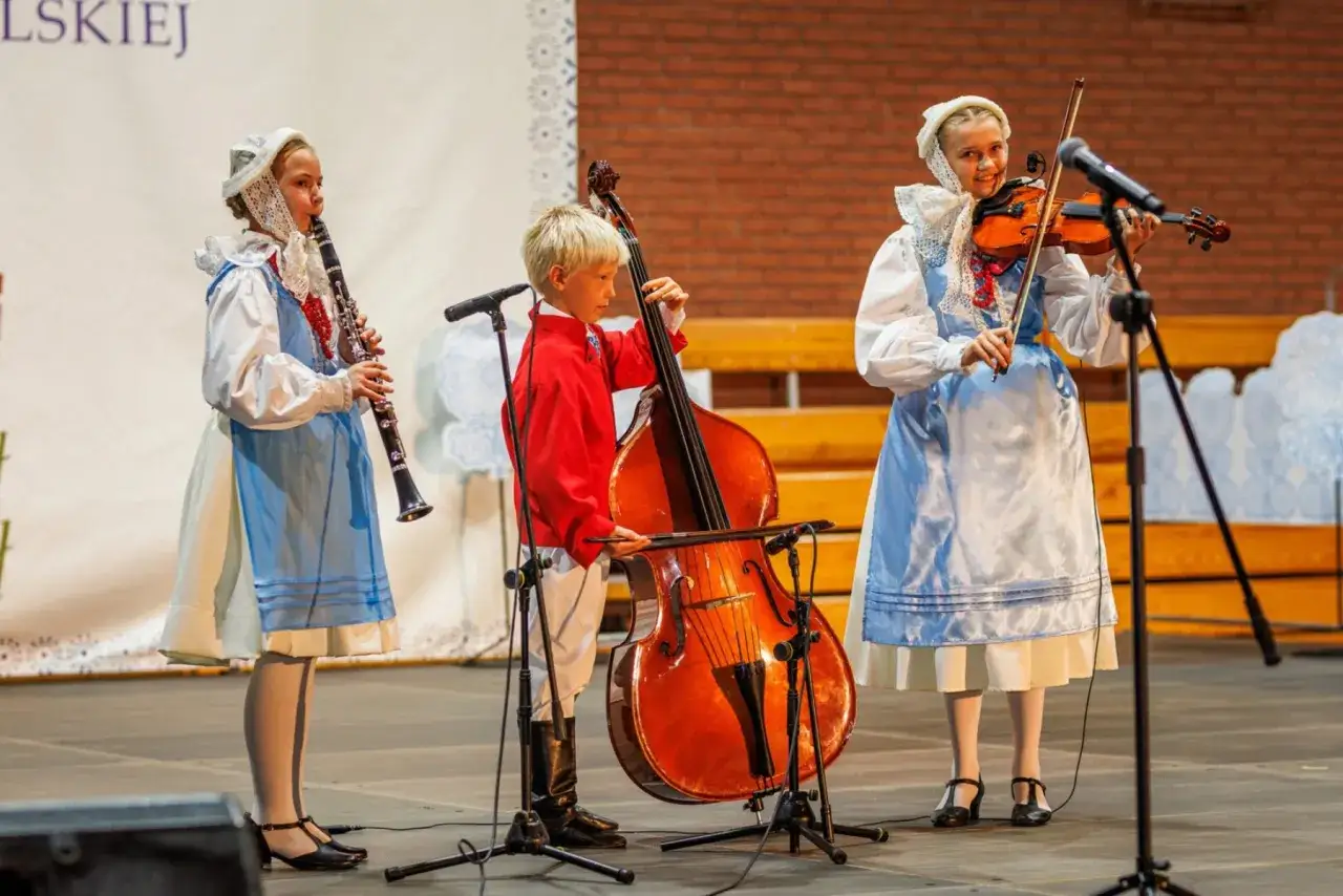 Dzieci w strojach ludowych grają na instrumentach: skrzypcach, kontrabasie i klarnecie. Chcą pokazać, że każdy może **zagraj to sam**!