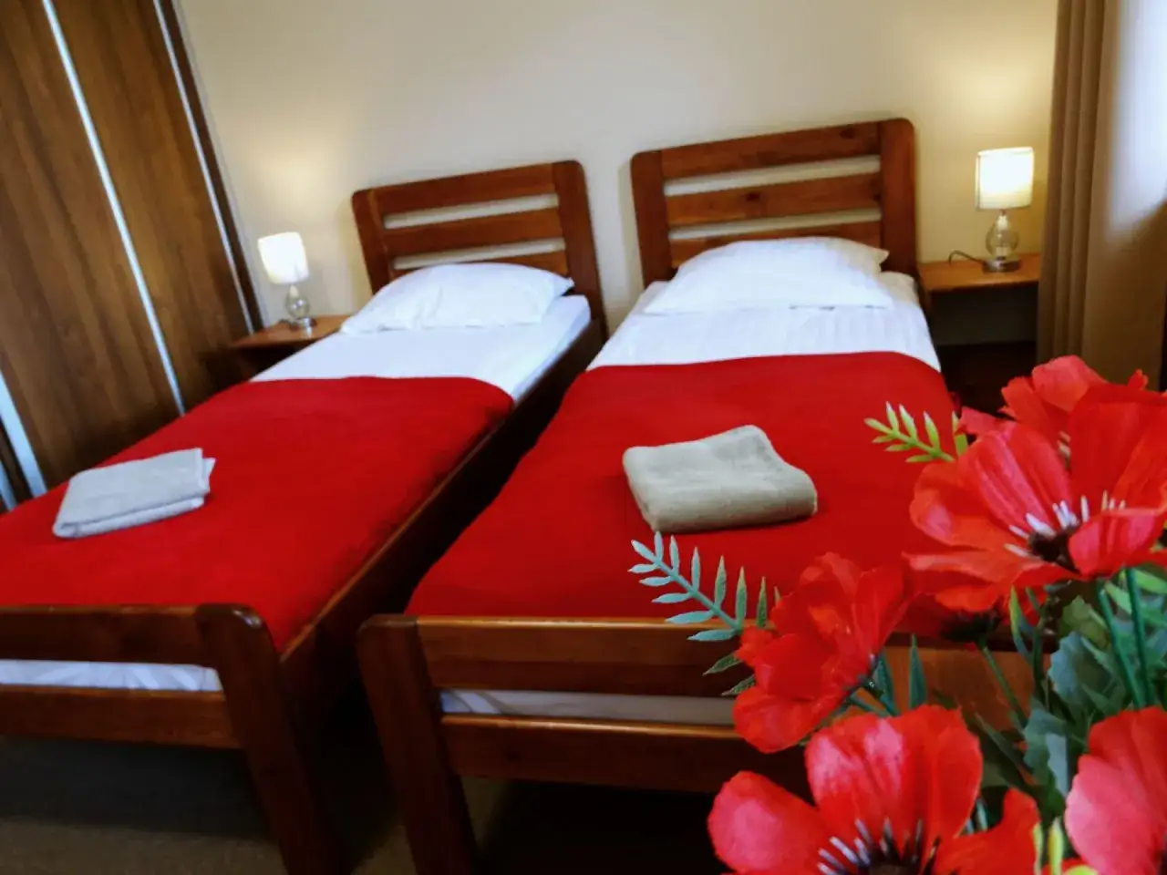 Dwa łóżka z czerwonymi narzutami i białymi pościelami w pokoju hotelowym. W rogu bukiet czerwonych maków. Idealne miejsce na wypoczynek w nowa fregata pokoje i apartamenty.