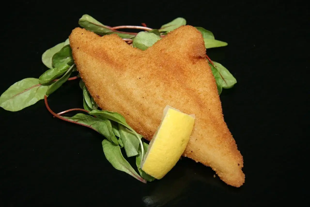 Gylla panerad fisk med en citronklyfta och gr&ouml;na blad p&aring; en svart tallrik.