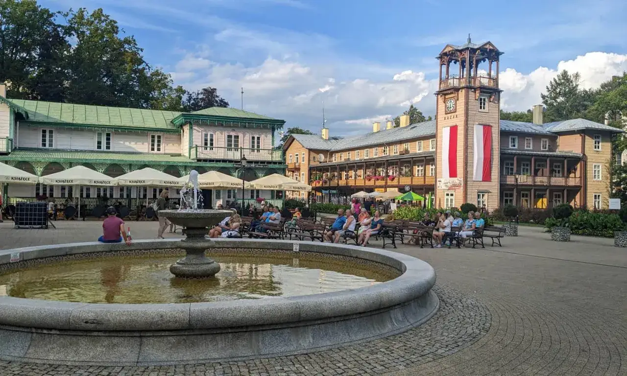 Rynek w Iwoniczu Zdroju z fontanną i ludźmi odpoczywającymi na ławkach. W tle zabytkowe budynki i wieża zegarowa. To jedne z głównych atrakcji Iwonicza Zdroju.