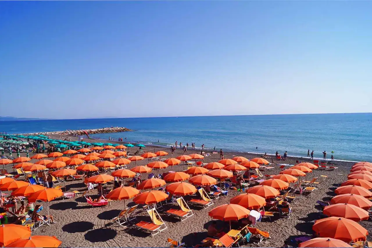 Spiaggia affollata con ombrelloni arancioni e verdi, lettini e persone che si godono il sole. Il mare azzurro e il cielo limpido creano una perfetta **sassetta distanza dal mare**.
