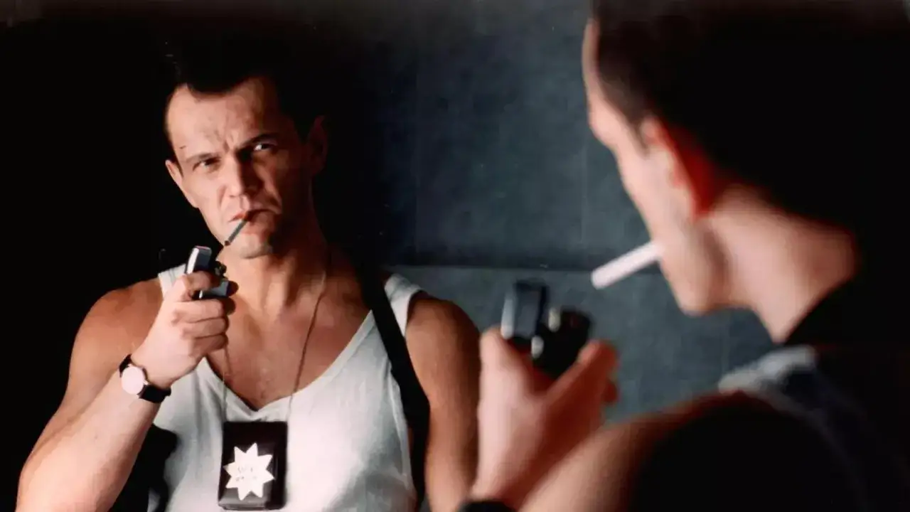 Mężczyzna z odznaką i papierosem, jak z polskich filmów z lat 90. W tle odbicie w lustrze.