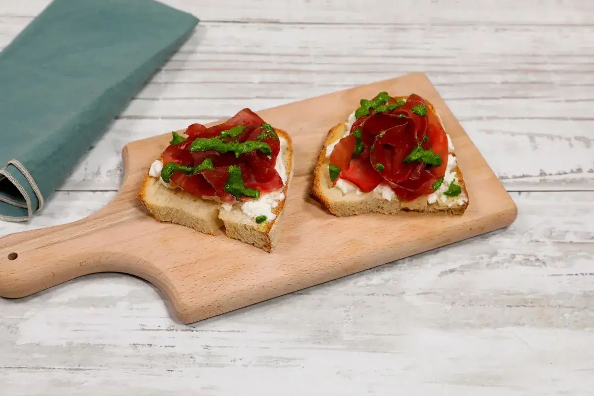 Bruschette con ricotta, bresaola e un tocco di pesto di rucola, su tagliere di legno.