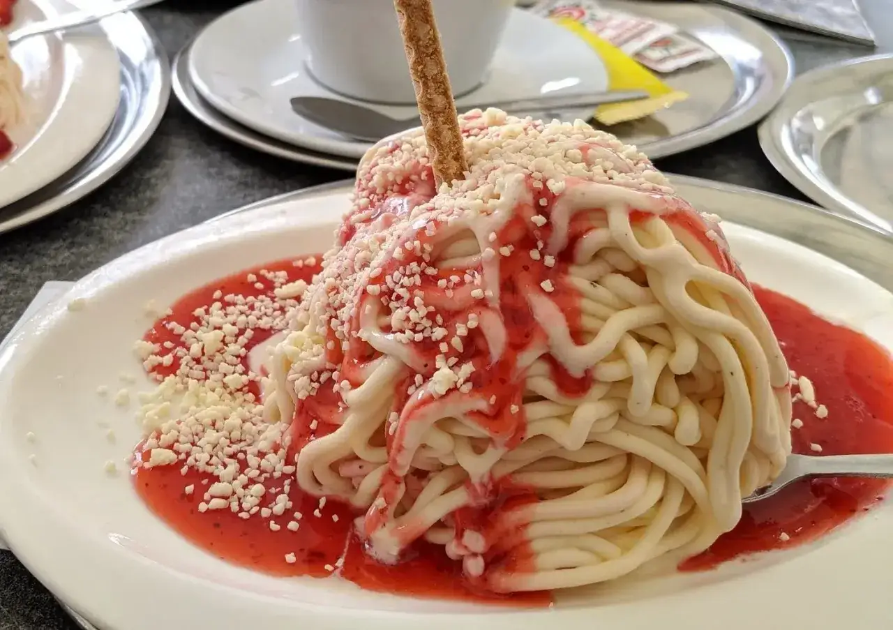 Ein s&uuml;&szlig;er Spaghetti-Kuchen auf einem Blech, verziert mit Erdbeersauce und wei&szlig;en Schokost&uuml;ckchen. Ein knuspriger Keks steckt obenauf.