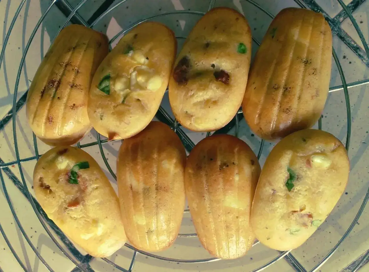 Huit madeleines salées, dorées et moelleuses, garnies de fromage et d'herbes, refroidissent sur une grille.