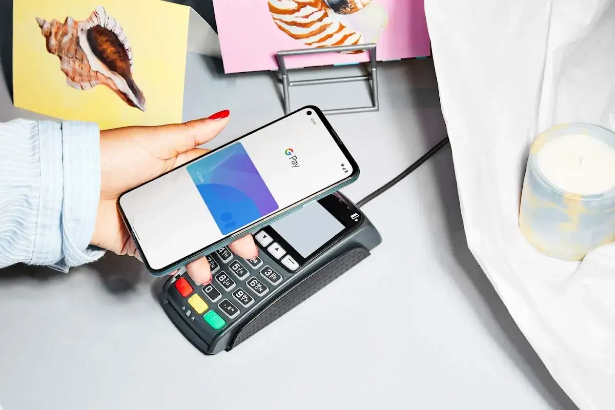 Dłoń zbliża telefon z otwartym portfelem Google Pay do terminala płatniczego.
