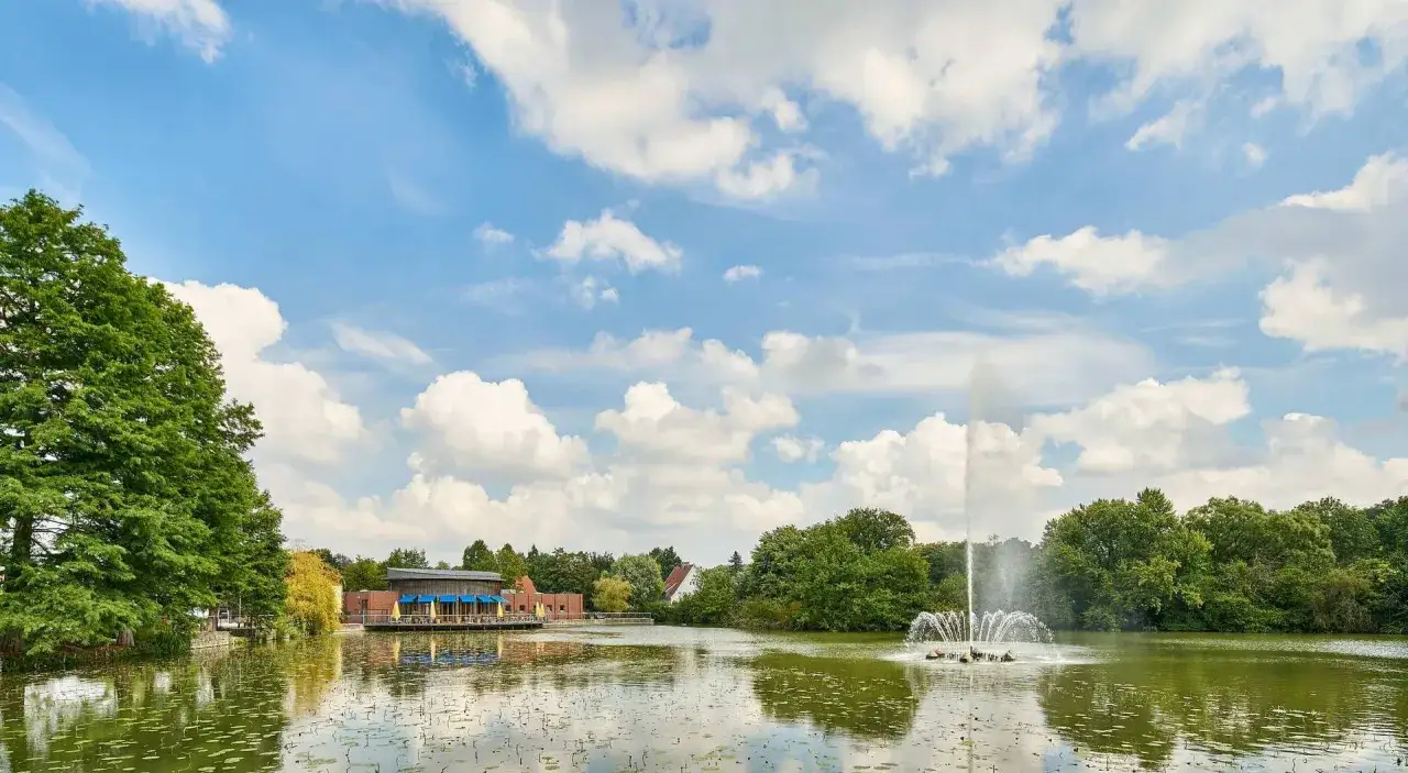 Ein See mit einem Springbrunnen im vier jahreszeiten park oelde. Im Hintergrund ein Gebäude mit blauen Sonnenschirmen.